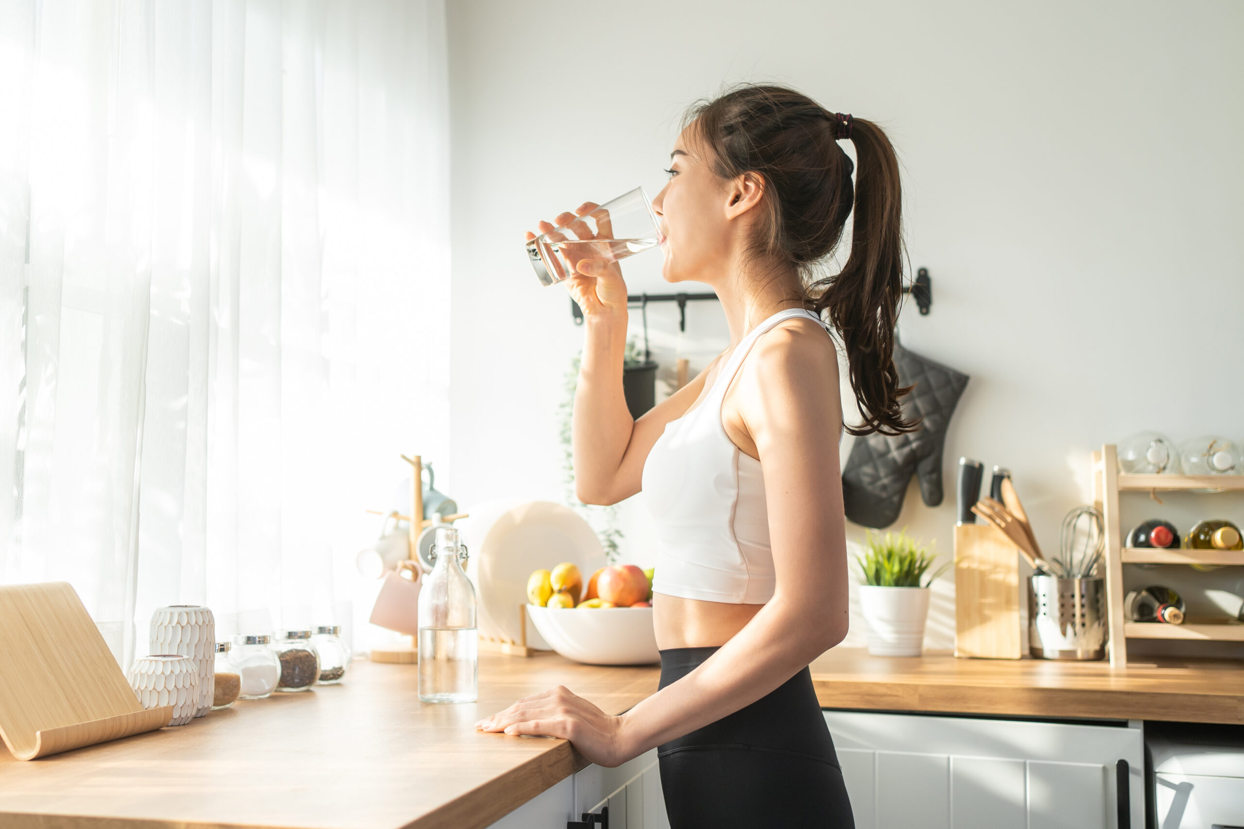 Asian beautiful woman in sportswear drink water after exercise at home. Young thirsty active sport girl takes a sips of clean mineral natural in cup after workout for health care in kitchen in house.