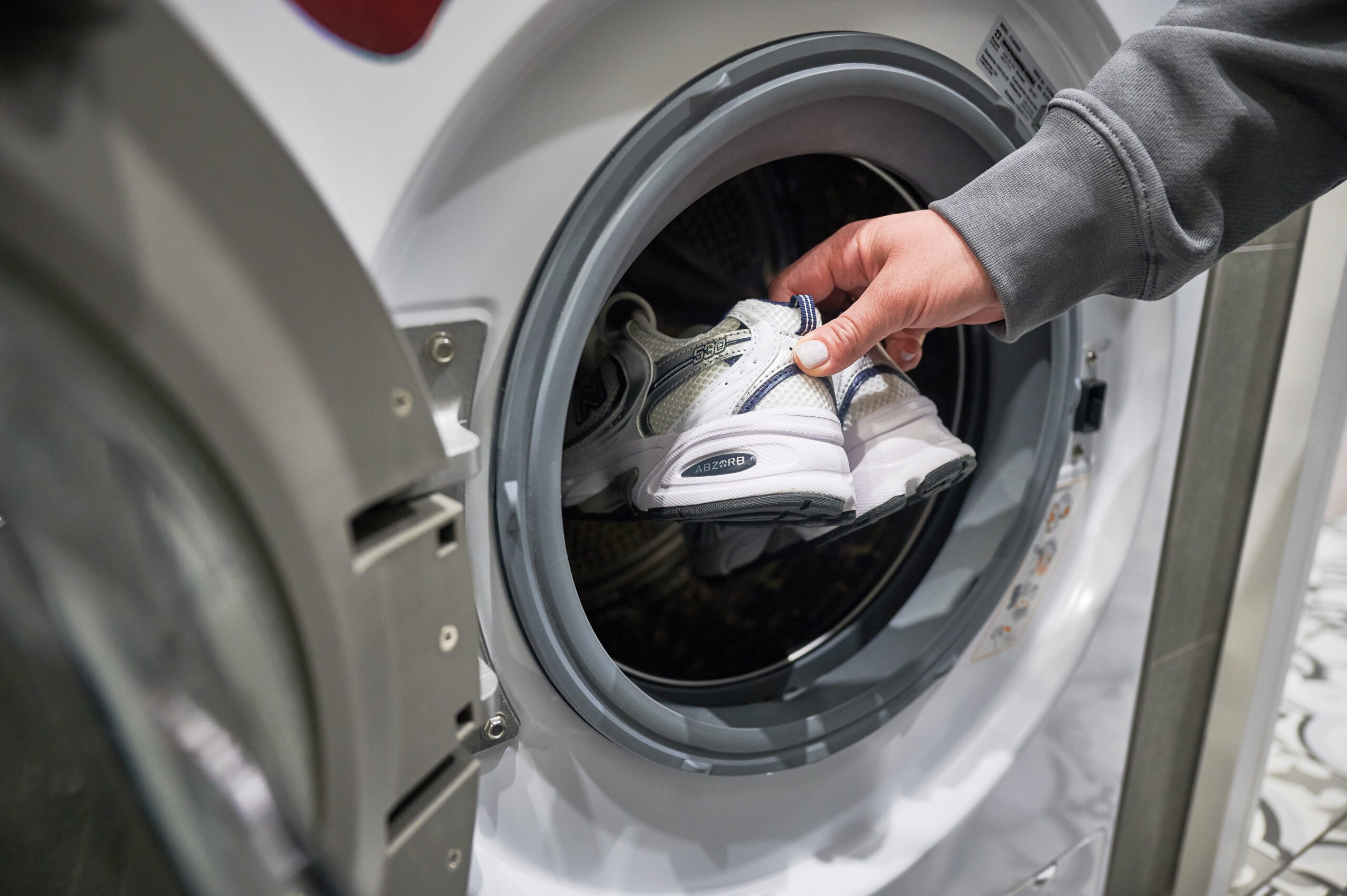 Lviv, Ukraine - May 7, 2023: Woman putting pair of sport shoes into washing machine. Washing white pair of New Balance 530 sneakers.